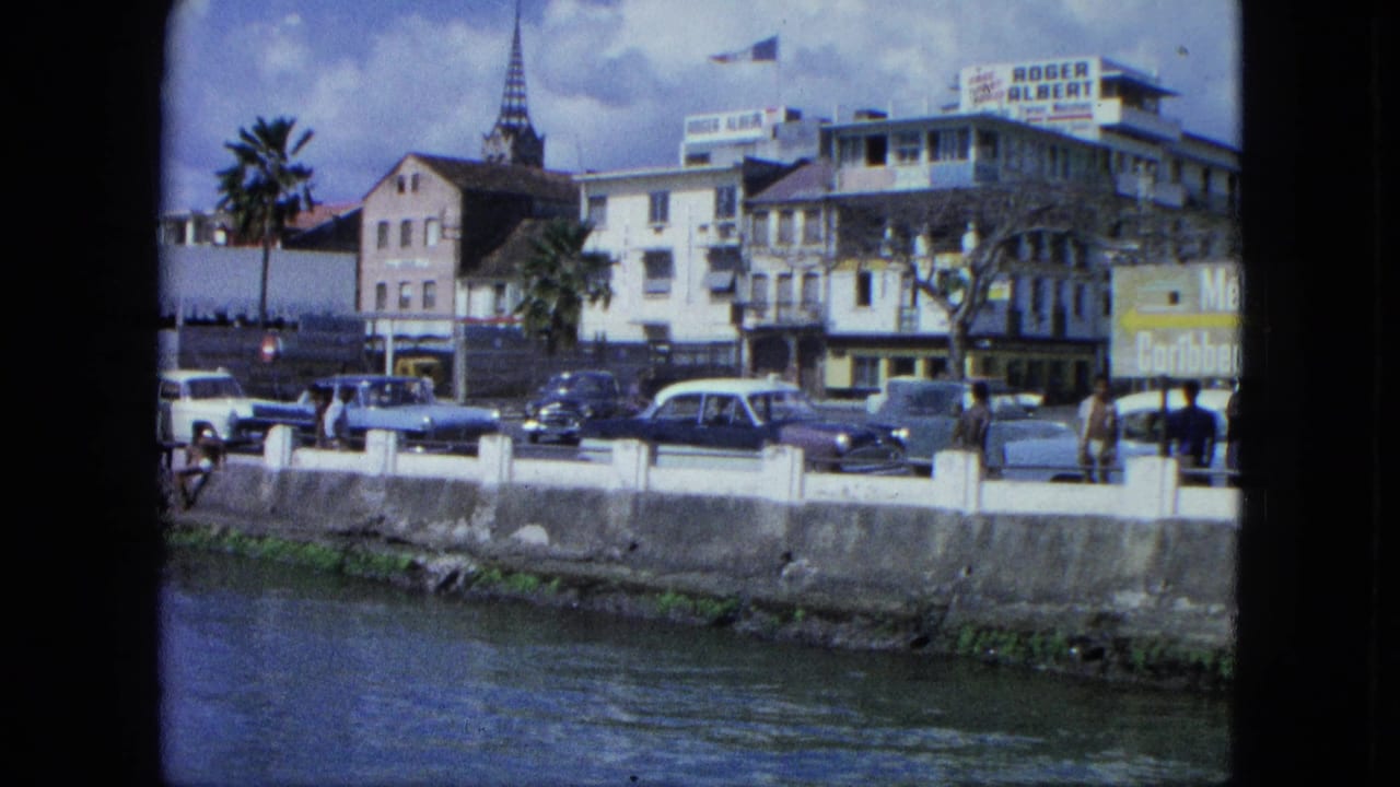 Waterfront street with mid-century cars, pedestrians, buildings, palm trees, and a church steeple.