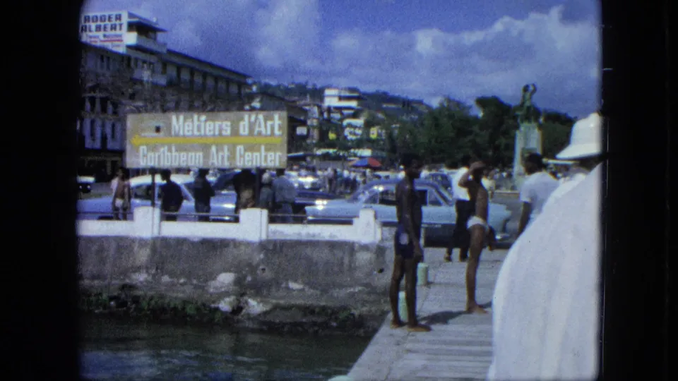 1964 USA Virgin Islands street scene with men by a canal, a Métiers d'Art Caribbean Art Center sign, parked cars, and hillside buildings.