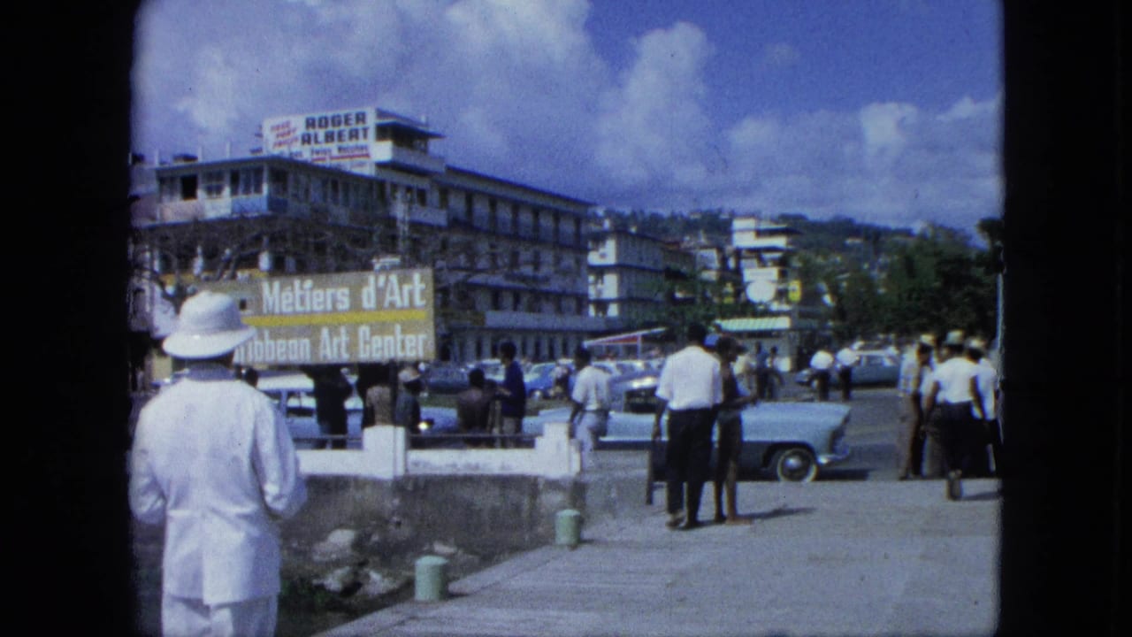 1964 USA Virgin Islands waterfront street view with a white-hatted man, classic cars, pedestrians, and visible art center and Roger Albert signs.