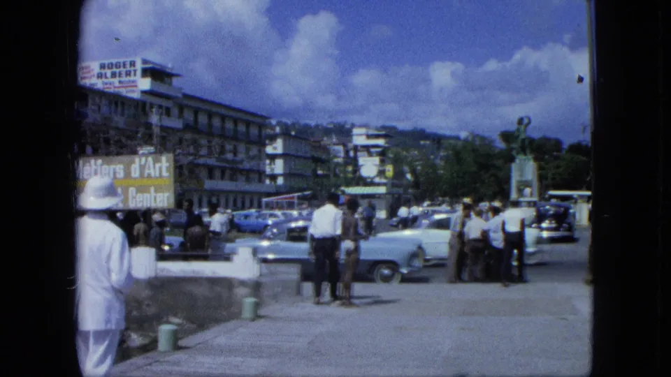 1964 USA Virgin Islands street scene with men on a concrete walkway, parked cars, commercial signs, and a statue near the road.