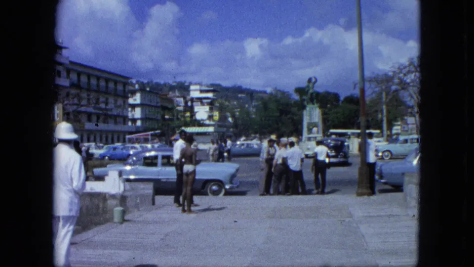 1964 USA Virgin Islands waterfront road with groups of men, classic cars, a statue, a pole, and hillside buildings under a bright sky.