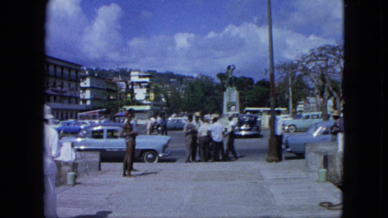 Men stand on a paved promenade near parked cars, a statue, buildings, and hillside greenery.