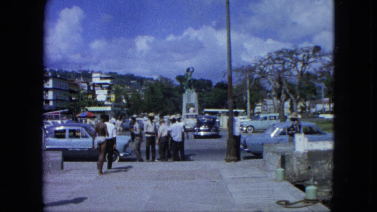 Pedestrians and parked cars line a waterfront promenade with a statue and town buildings behind them.