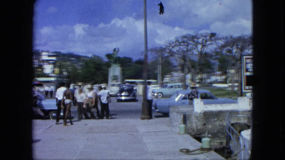 1964 8mm frame of a Caribbean public promenade with men near parked cars, a pedestal statue, a lamppost, and a visible welcome sign.