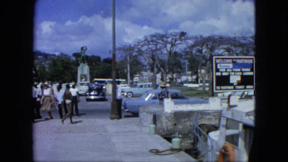 1964 8mm frame showing pedestrians crossing a Caribbean promenade near classic cars, a monument, and a welcome sign by the waterfront.
