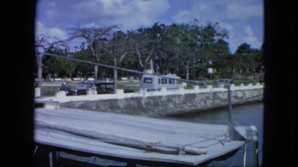 1964 USA Virgin Islands marina frame with dock rigging, a seawall, classic cars, a light VW camper-style van, trees, and a visible person.