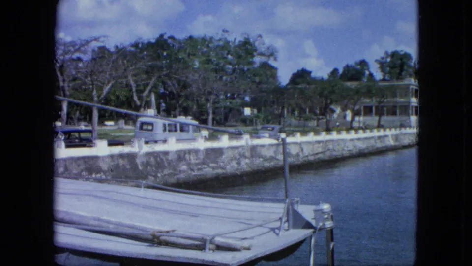 1964 USA Virgin Islands waterfront view with marina water, dock edge, stone seawall, light VW camper-style van, parked cars, trees, and a building.