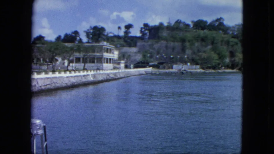 1964 U.S. Virgin Islands harbor view with broad blue water, a stone seawall, a white porch-front house, roadside area, and dense tropical vegetation.