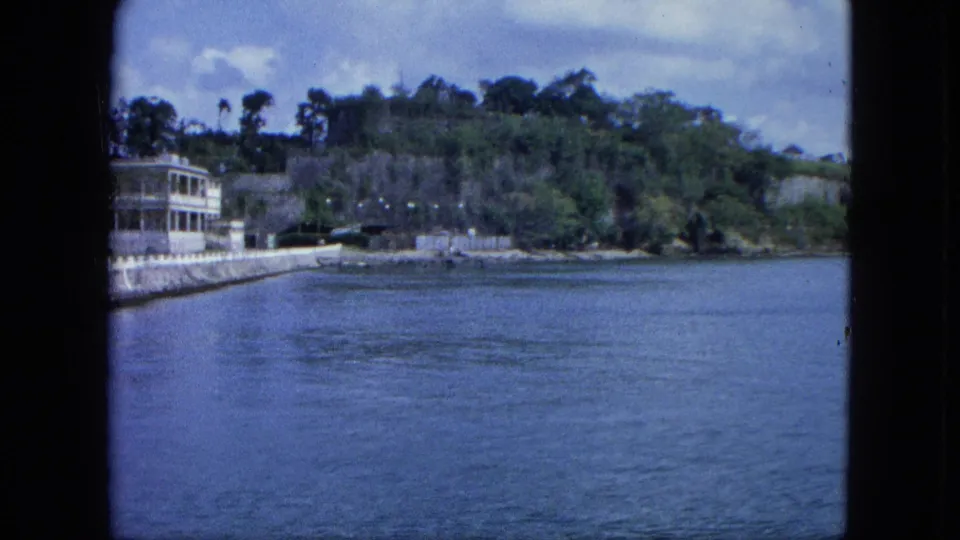 1964 U.S. Virgin Islands harbor scene with open water, shoreline seawall, partial white porch house, parked vehicles, and tropical hillside vegetation.