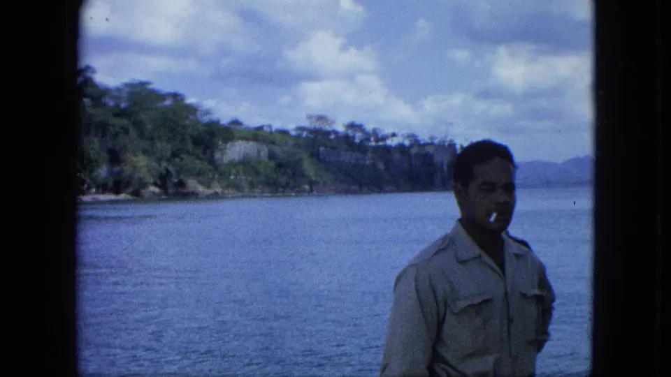 Adult man in a light shirt smokes by a window while standing before a 1964 U.S. Virgin Islands harbor and wooded shoreline.