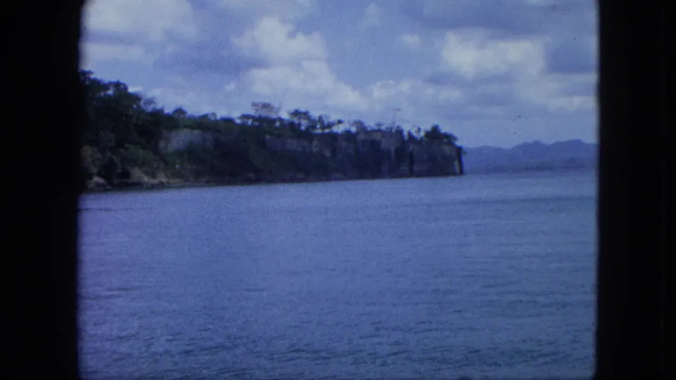 1964 8mm U.S. Virgin Islands view across calm harbor water toward a wooded headland, cloudy sky, and distant hills.