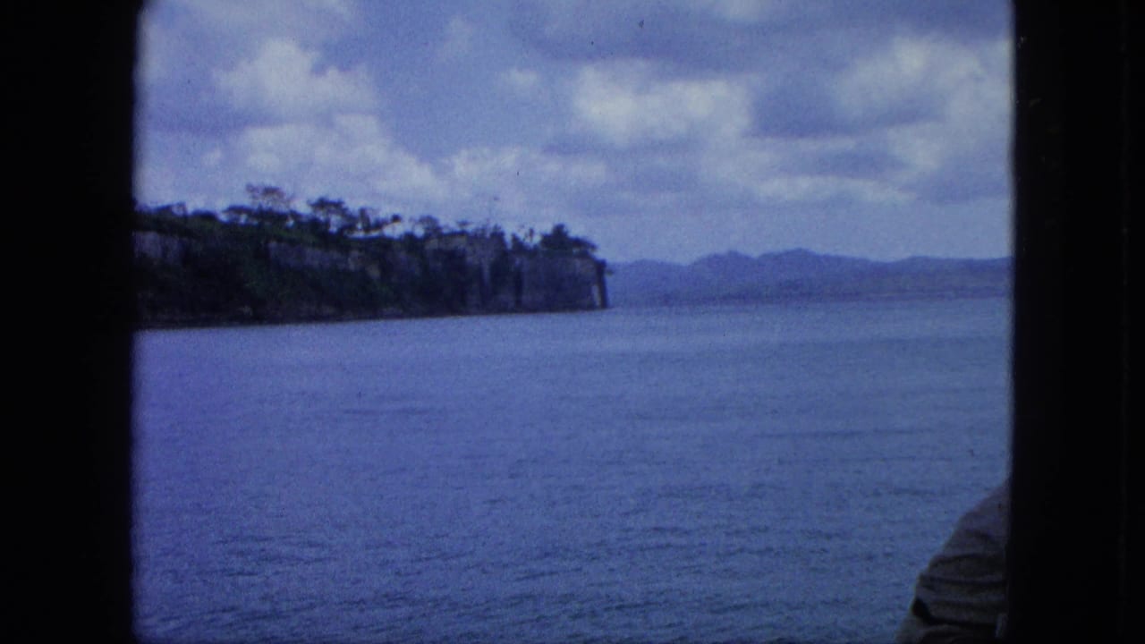 Coastal water view with cliffs, distant hills, and part of a passenger at right.