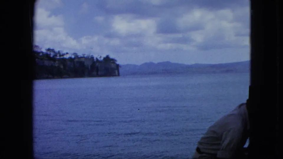 1964 U.S. Virgin Islands 8mm frame with calm sea, distant shore, clouded sky, and a partial adult man in a light shirt at lower right.