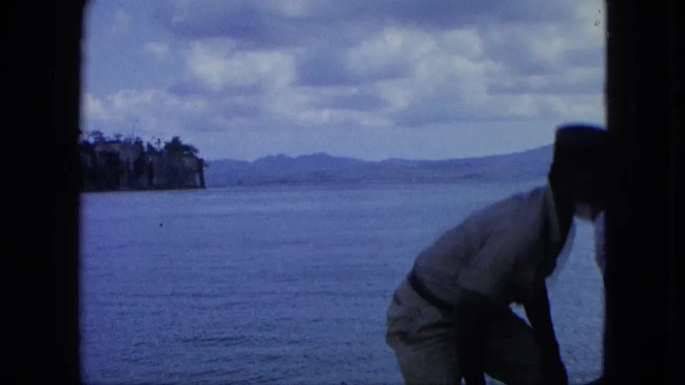 1964 U.S. Virgin Islands boat scene with an adult man in a dark hat foregrounded against blue sea, cliffs, distant hills, and clouds.