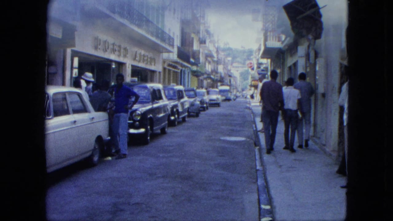 Narrow street with parked cars, pedestrians, storefronts, and balconies.