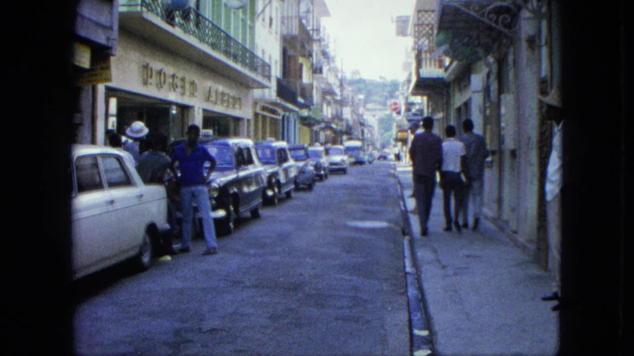 Men walk along a sidewalk beside parked cars on a narrow street.