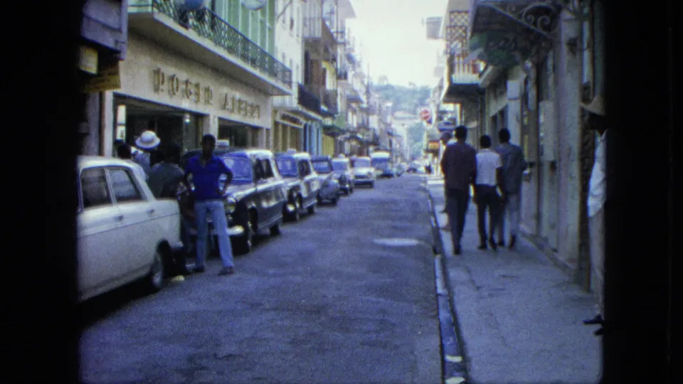 1964 USA Virgin Islands street scene with men walking away from the camera, parked sedans, shopfronts, and upper-floor balconies.