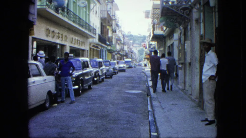 1964 USA Virgin Islands street view showing a man near a parked car, pedestrians ahead, storefront signage, balconies, and a narrow roadway.