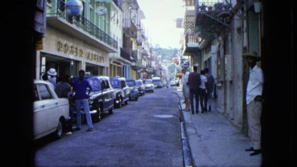 1964 USA Virgin Islands street scene with pedestrians on the sidewalk, a row of parked classic cars, storefront signage, and balcony railings.