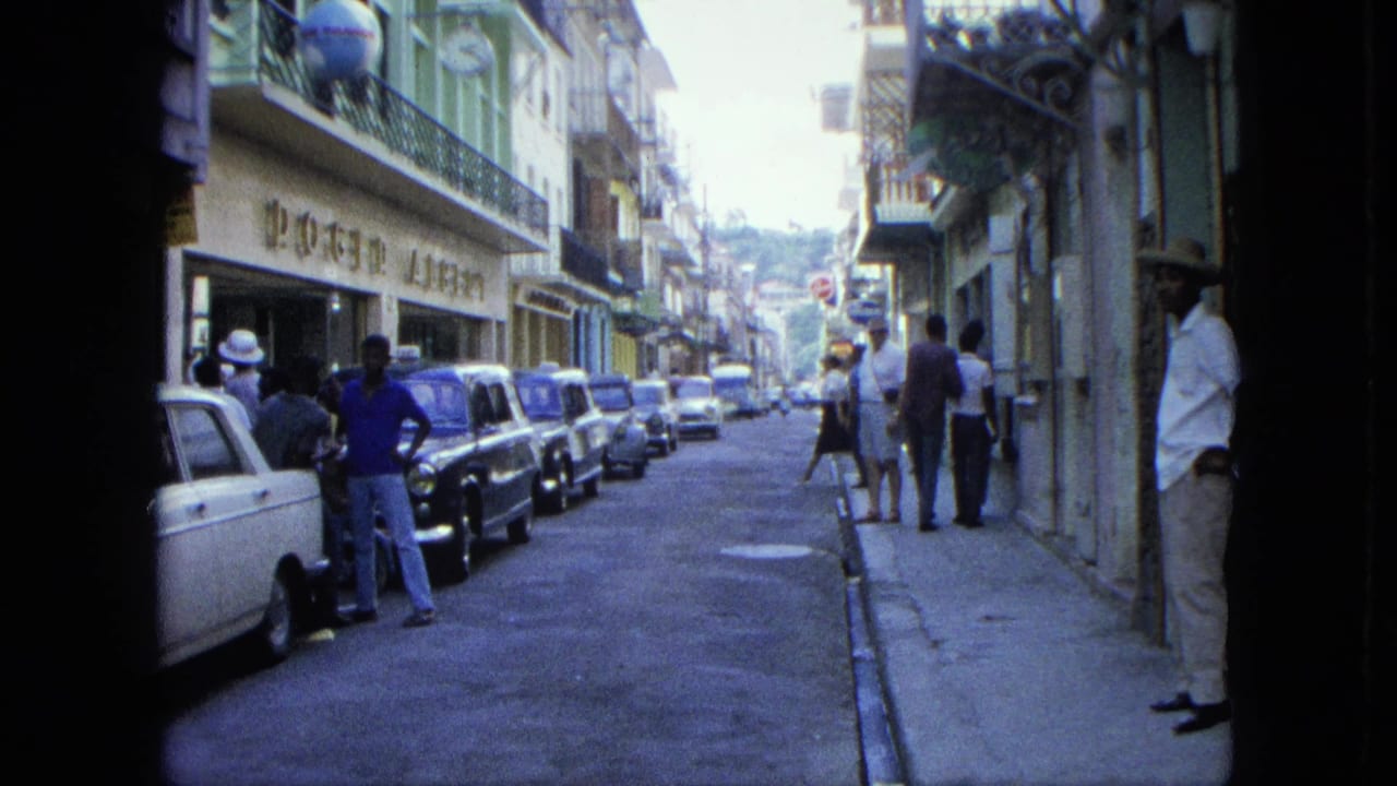 Narrow street with parked cars, pedestrians, storefronts, balconies, and hillside buildings in the distance.