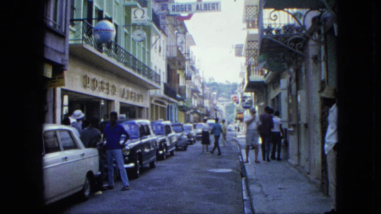 1964 U.S. Virgin Islands street view showing pedestrians near shopfronts, parked mid-century cars, upper balconies, and signage along the road.