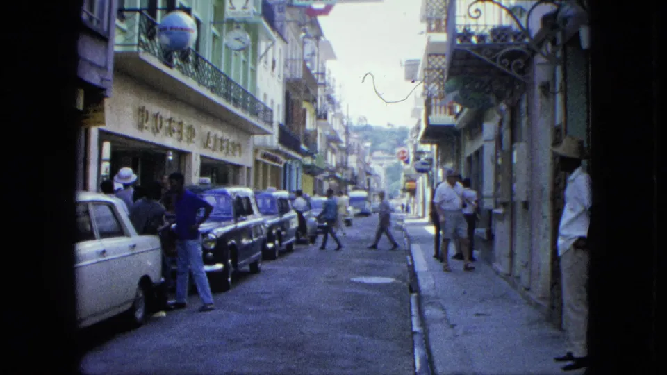 1964 U.S. Virgin Islands frame with pedestrians crossing and standing beside parked classic cars along a shop-lined street.