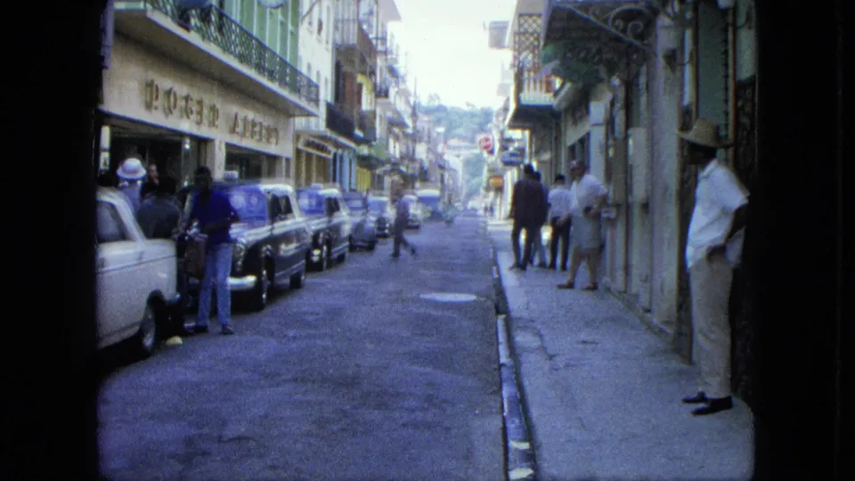 1964 U.S. Virgin Islands home-movie frame of a shaded shop street with parked cars, pedestrians, signs, balconies, and film gate edges visible.