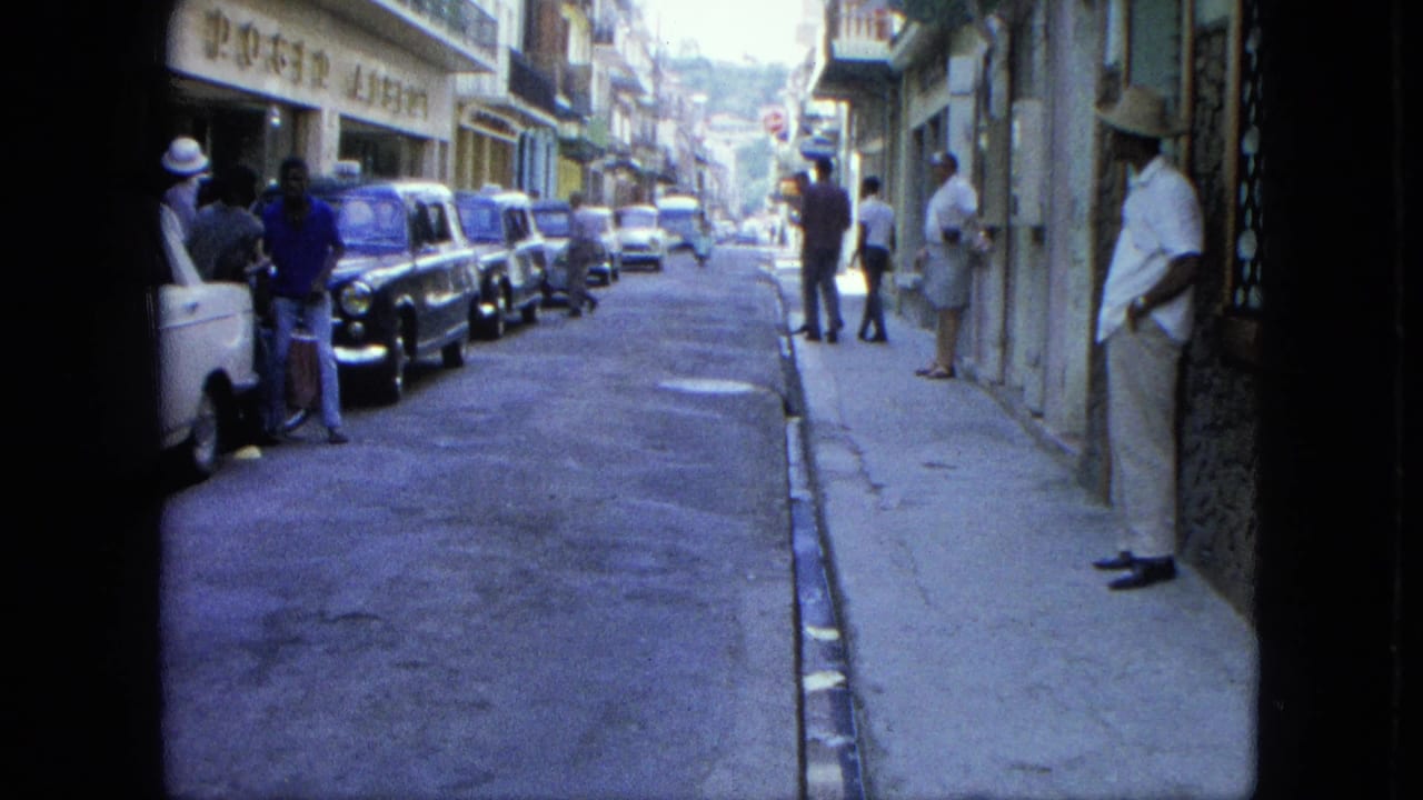 Narrow street with parked cars, pedestrians, storefronts, and a distant hillside.