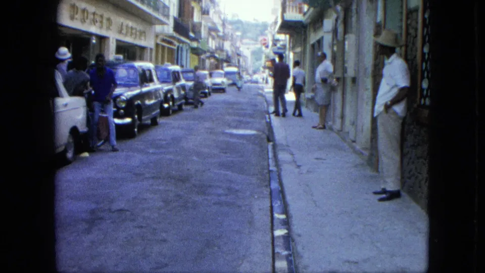 1964 U.S. Virgin Islands street view with parked mid-century cars, pedestrians near storefronts, and a sloping road in bright daylight.