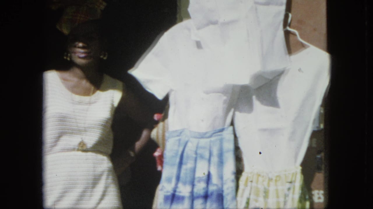 Woman in a white dress and headwrap stands beside hanging shirts and skirts.