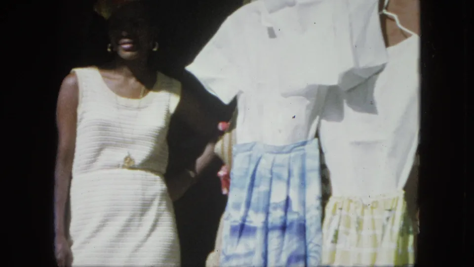 Closer 8mm view of the woman in a white dress standing next to hanging white shirts and blue and yellow skirts outside a shop.