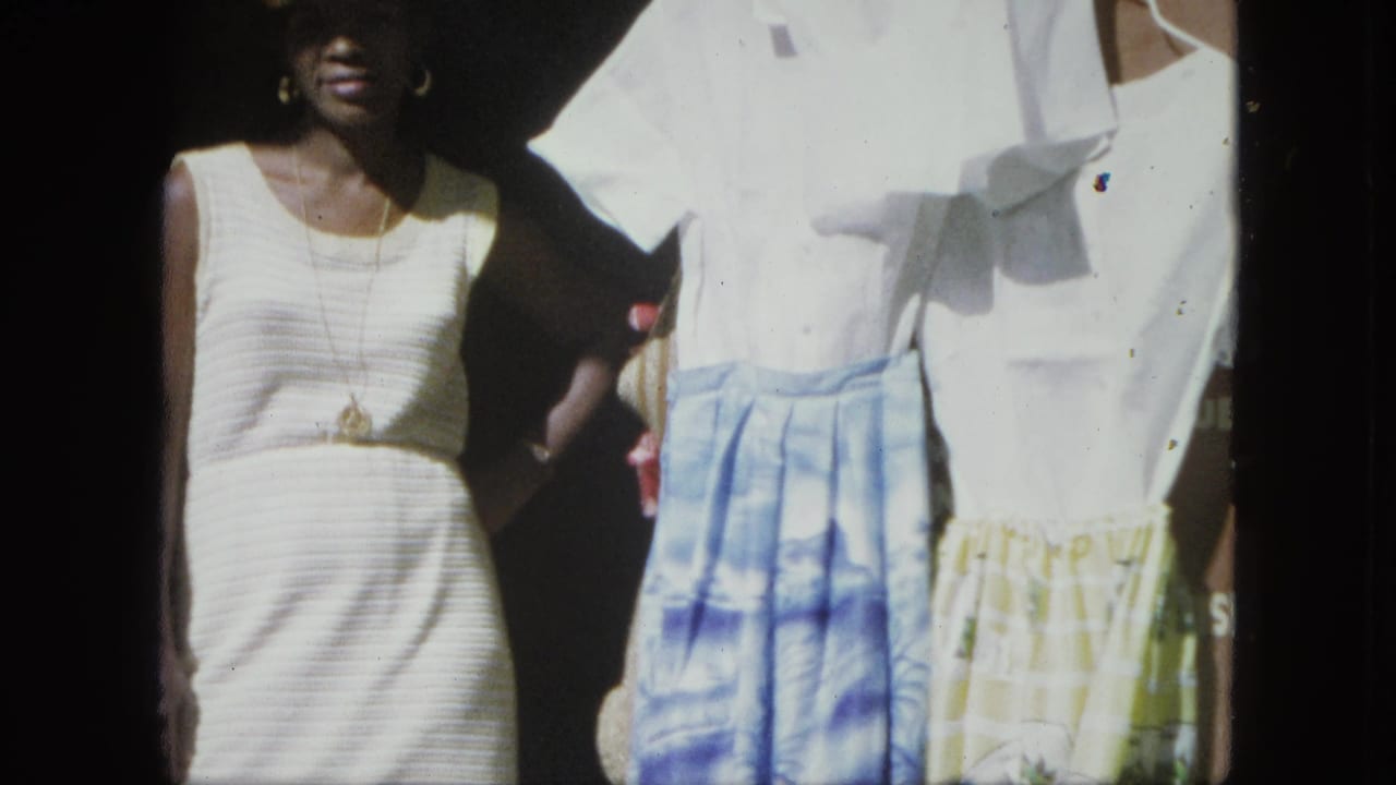 In 1964 USA Virgin Islands home-movie footage, a woman in a white dress stands beside blue and yellow patterned skirts.
