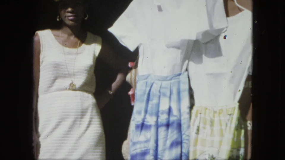 In 1964 USA Virgin Islands home-movie footage, a woman in a white dress stands beside blue and yellow patterned skirts.