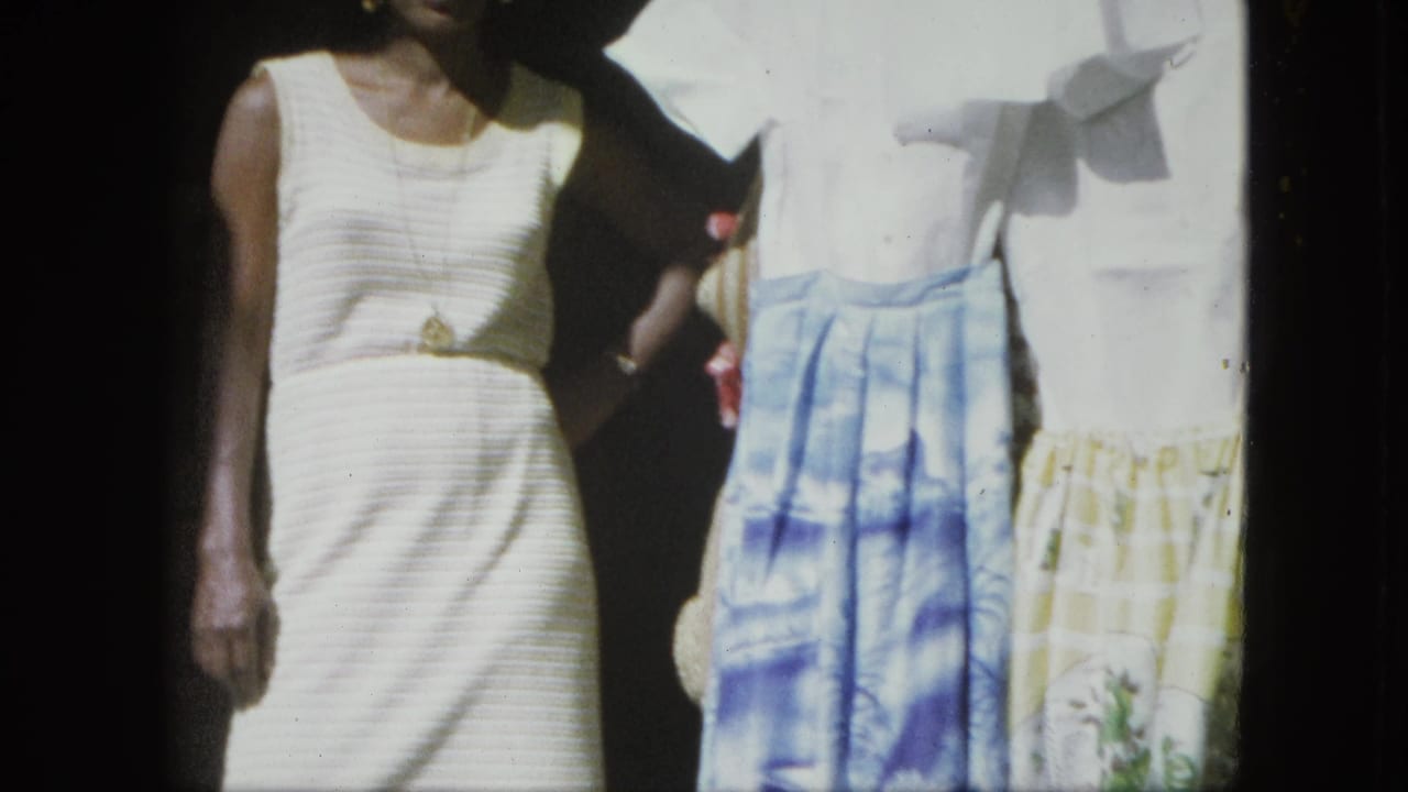 The woman remains beside a clothing display, with ribbed white dress, necklace, and patterned skirts visible in the frame.