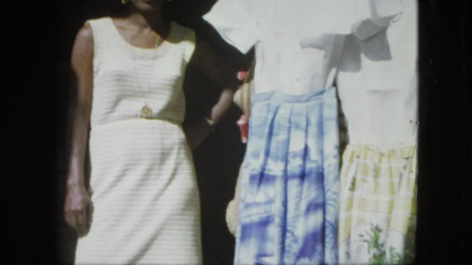 The woman remains beside a clothing display, with ribbed white dress, necklace, and patterned skirts visible in the frame.