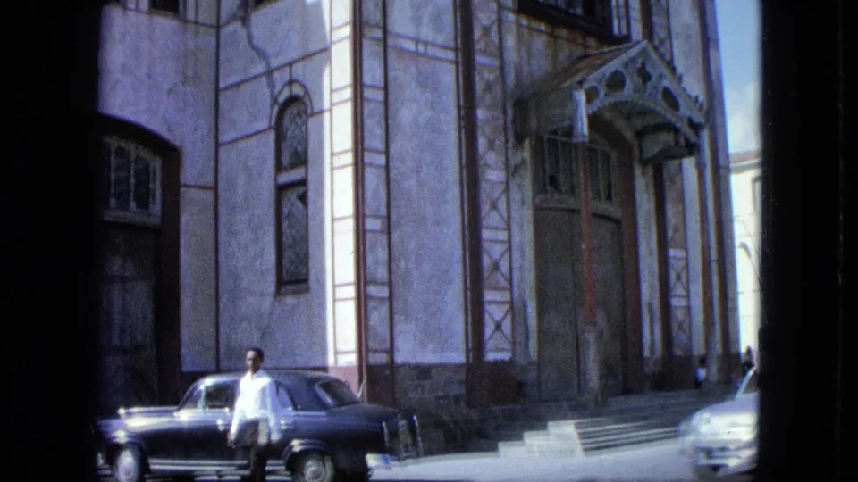 A street exterior shows a man near a parked car outside a church-like facade with steps and tall windows.