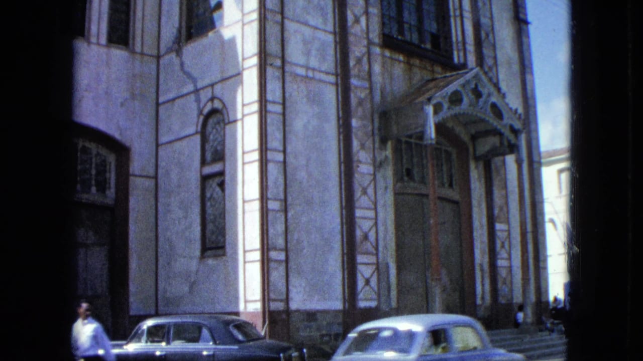 Ornate light-colored building facade with arched windows, parked cars, and a person walking nearby.