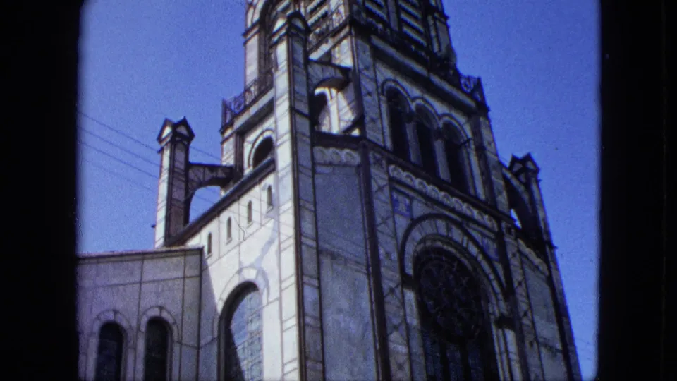 1964 8mm frame showing a low-angle exterior view of a pale church tower, arched stained glass, roofline details, and overhead wires.