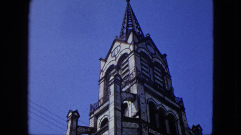 1964 8mm frame showing the church spire and upper tower from below, with roofline masonry, arched details, and visible film grain.