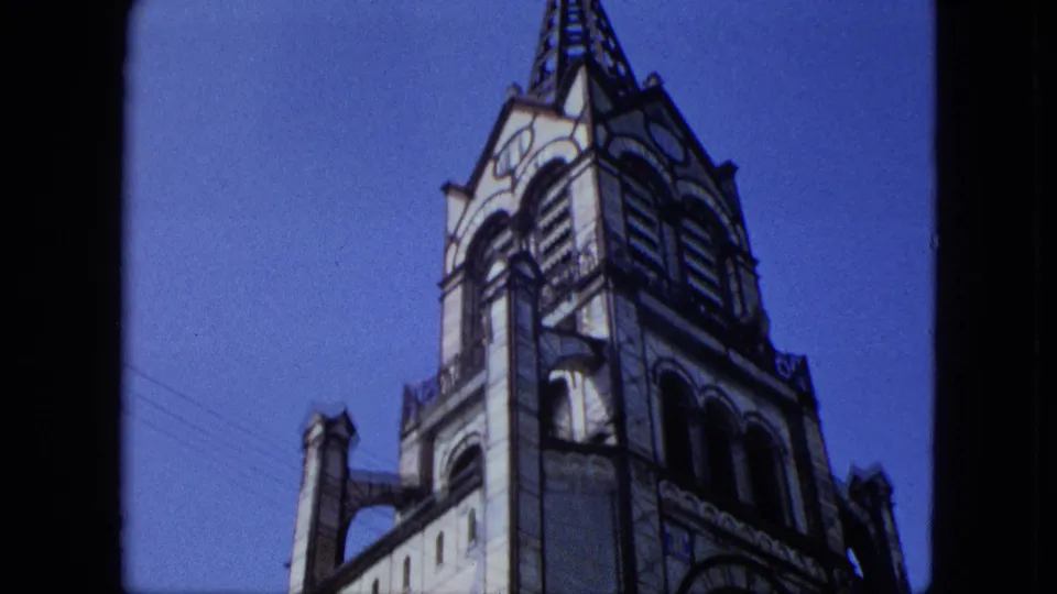 1964 8mm frame of the ornate church tower and spire, with arched facade details, a side roofline, power cables, and clear sky.