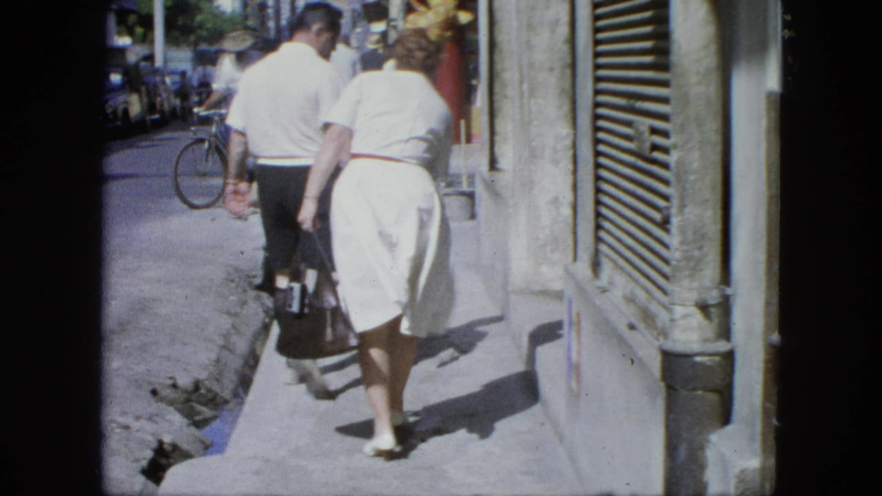 1964 United States home-movie frame of an elderly couple walking away on a sunny sidewalk, with street traffic and a bicycle behind them.