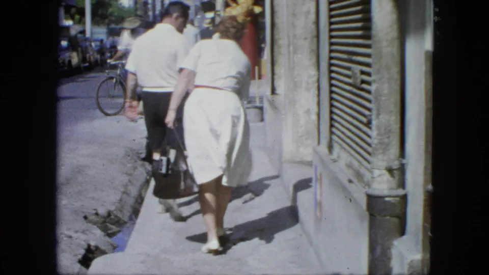 1964 United States home-movie frame of an elderly couple walking away on a sunny sidewalk, with street traffic and a bicycle behind them.
