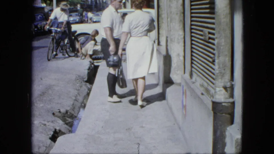 1964 United States street scene with the couple walking away, a cyclist nearby, parked mid-century cars, and a shuttered shopfront.
