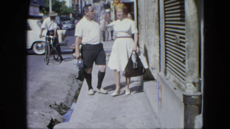 1964 United States home-movie frame showing the couple facing the camera near a storefront, with a cyclist and classic cars behind them.