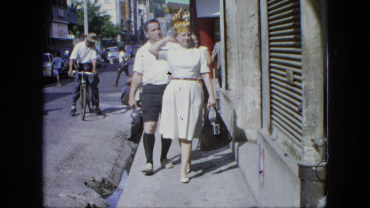 Couple walking toward the camera on a sunny sidewalk with pedestrians and cars behind them.
