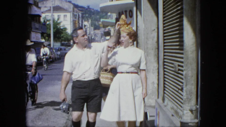 1964 U.S. Virgin Islands home-movie frame showing the woman gesturing near her headpiece as the couple walks past a storefront.