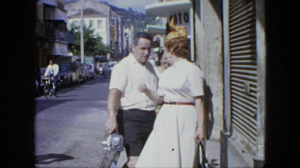 1964 U.S. Virgin Islands frame showing the couple close together on the sidewalk, with the man holding a small camera and cars behind them.