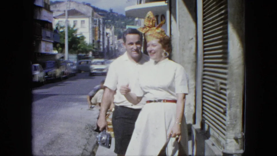 In 1964 U.S. Virgin Islands home-movie footage, a man and a smiling woman in a white dress walk along a busy street.