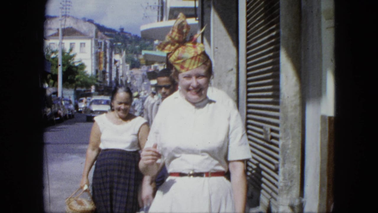 A smiling woman in a white dress and yellow-red headscarf walks toward the camera on a U.S. Virgin Islands street in 1964 footage.