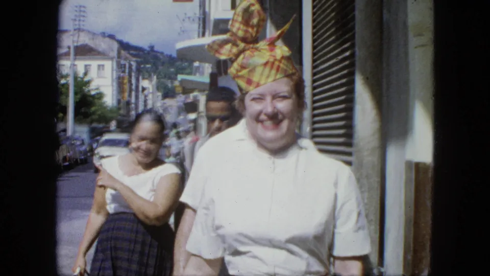 The woman in a patterned headscarf moves nearer to the camera while other adults walk behind her on a sunny urban sidewalk.